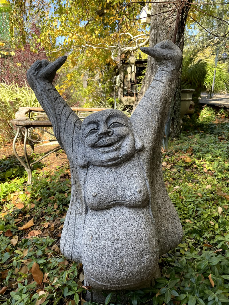 Granite buddha in garden setting, happy face, round tummy, hands in air.